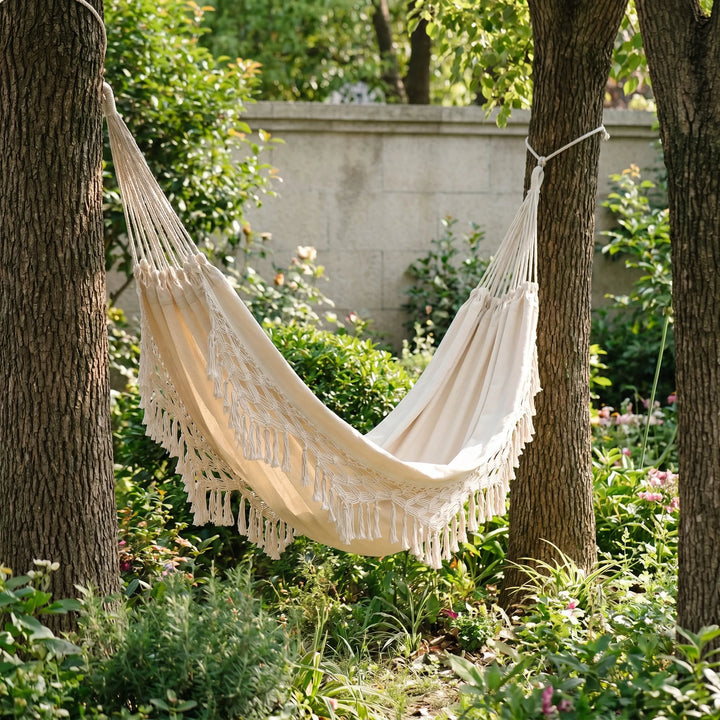 Boho tasseled cotton outdoor hammock hanging in a lush garden, perfect for relaxation & bohemian living space.