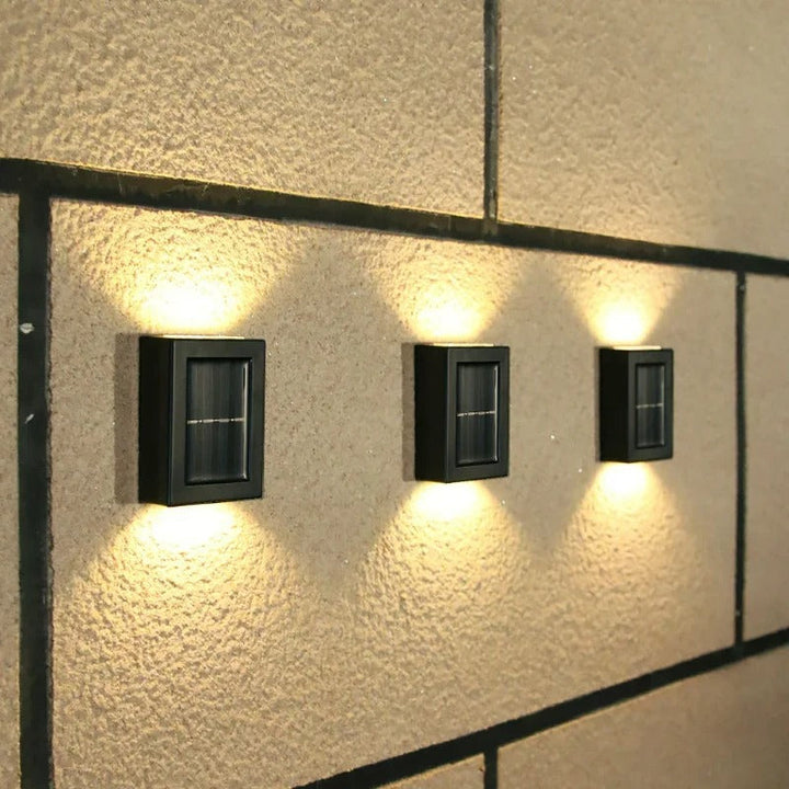 Sleek black outdoor solar LED wall fixture casting a warm glow on a garden pathway at dusk.