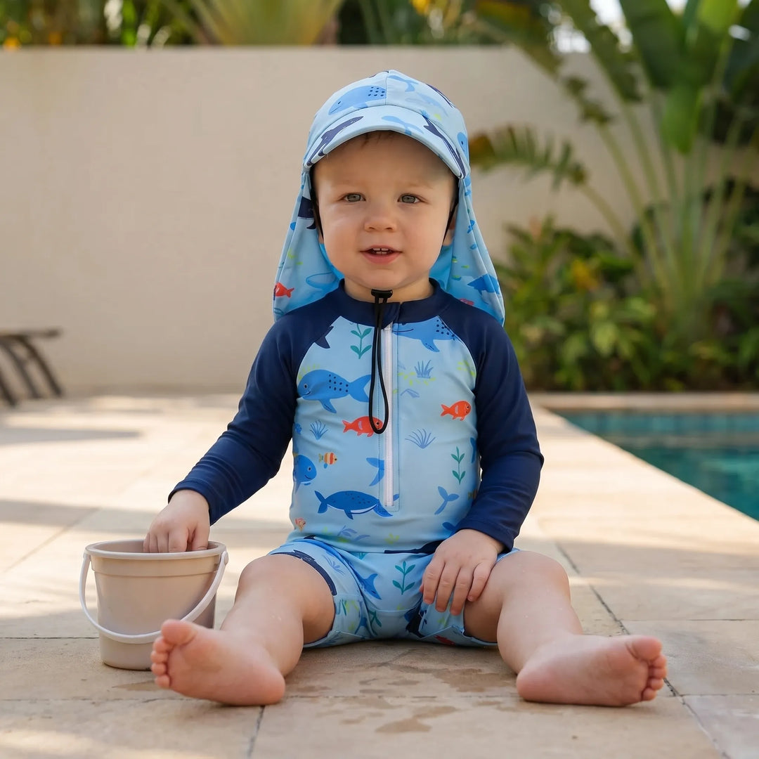 Toddler in blue marine print long-sleeve swim romper & sun hat. Enjoying water play at the beach with UV protection.